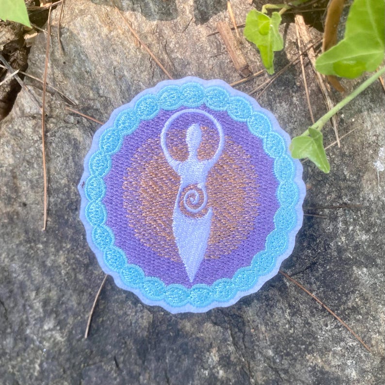 May include: Embroidered patch with a white figure in a purple and gold circular design, bordered by a light blue scalloped edge. The patch is set on a grey stone surface with green foliage. The design depicts a stylized female form.