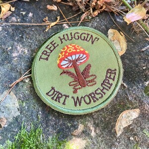 Pu&ograve; includere: Toppa rotonda verde oliva con testo ricamato e design a fungo. Il testo recita "TREE HUGGIN'" e "DIRT WORSHIPER". Il fungo ha un cappello rosso e arancione, lamelle bianche e un gambo marrone. Su una superficie strutturata.