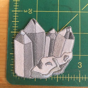 May include: Embroidered patch depicting a cluster of crystals in shades of grey and white. The patch has a detailed design with various facets and textures, resembling a geode. The patch is placed on a green cutting mat with measurement markings.