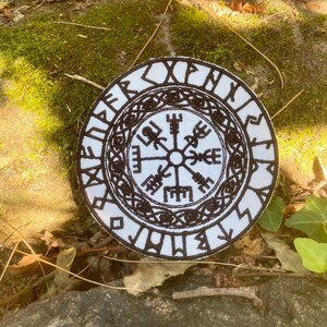 May include: A circular white patch with a detailed black Viking symbol design. The patch has runes around the border and a central compass-like emblem. The design is intricate and looks embroidered or stitched.