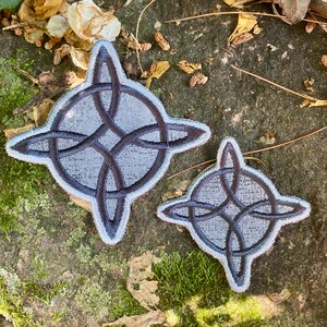 May include: Two embroidered patches featuring a Celtic knot design. The patches are gray with dark gray knotwork and a light gray border. One patch is larger than the other. The patches are displayed on a textured, natural surface.