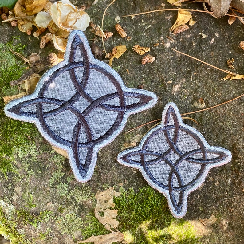 May include: Two embroidered patches featuring a Celtic knot design. The patches are gray with dark gray knotwork and a light gray border. One patch is larger than the other. The patches are displayed on a textured, natural surface.