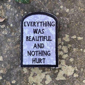 May include: A rectangular patch with rounded top edges, featuring the text "EVERYTHING WAS BEAUTIFUL AND NOTHING HURT" in black on a light purple background. The patch has a black border and is likely an iron-on or sew-on patch.