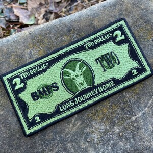 May include: A rectangular patch resembling a two-dollar bill, featuring a green and black color scheme. The design includes a goat head emblem, the word "TWO" in large letters, and the text "LONG JOURNEY HOME."