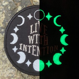 May include: Circular black patch featuring white embroidered moons, stars, and the text "LIVE WITH INTENTION." The right side glows green in the dark, displaying the same celestial design. A unique, mystical accessory.