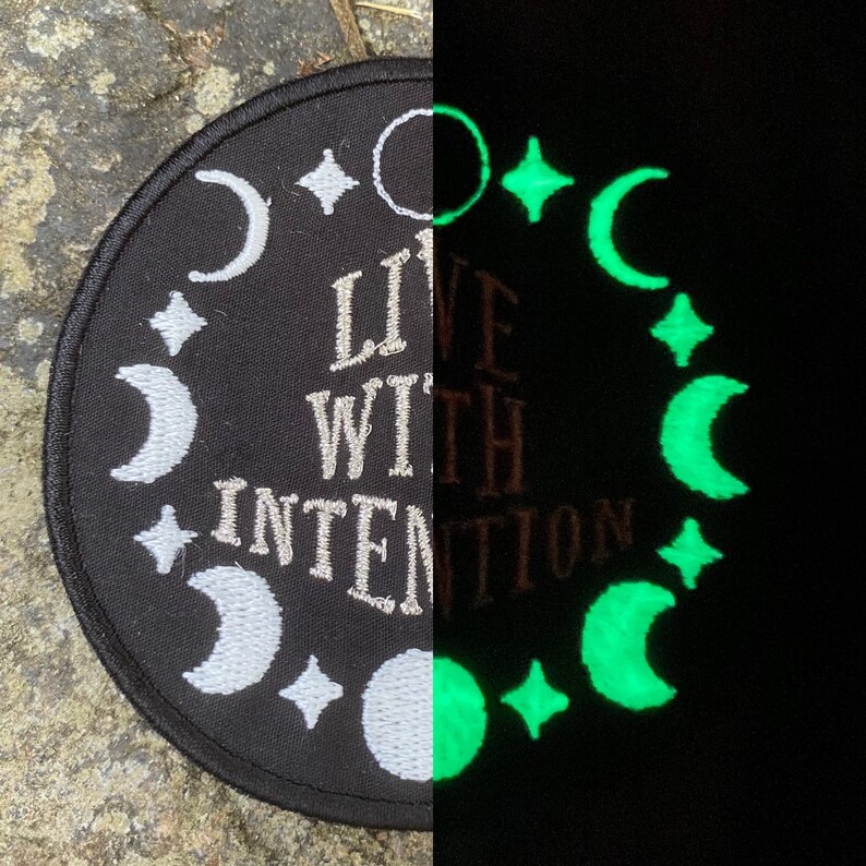 May include: Circular black patch featuring white embroidered moons, stars, and the text "LIVE WITH INTENTION." The right side glows green in the dark, displaying the same celestial design. A unique, mystical accessory.