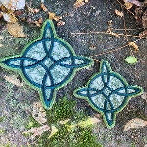 May include: Two Celtic knot patches. The larger patch is approximately 4 inches in diameter, and the smaller one is about 2.5 inches. Both have a green and blue design with a floral pattern in the center. The patches are on a textured, natural surface.