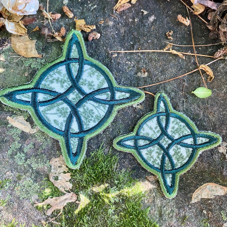 May include: Two Celtic knot patches. The larger patch is approximately 4 inches in diameter, and the smaller one is about 2.5 inches. Both have a green and blue design with a floral pattern in the center. The patches are on a textured, natural surface.