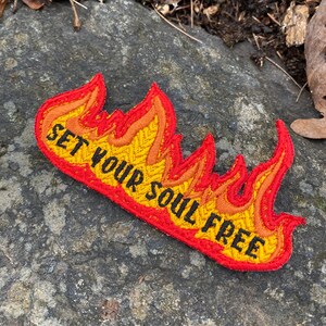 May include: Embroidered patch in the shape of flames, with a red border and yellow, orange, and red interior. The text "SET YOUR SOUL FREE" is embroidered in black across the center of the flames.