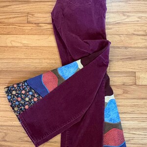 Op de afbeelding: Pruimkleurige corduroy broek met een decoratief patchwork ontwerp op de onderbenen. Het patchwork heeft blauwe, rode en bruine vormen en een bloemenpatroon. De broek ligt op een houten oppervlak.