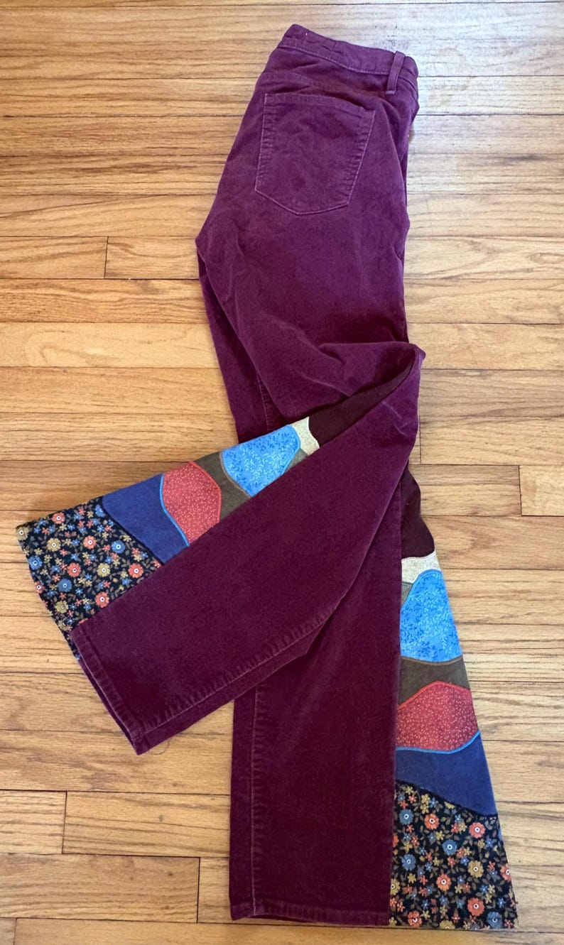 Op de afbeelding: Pruimkleurige corduroy broek met een decoratief patchwork ontwerp op de onderbenen. Het patchwork heeft blauwe, rode en bruine vormen en een bloemenpatroon. De broek ligt op een houten oppervlak.