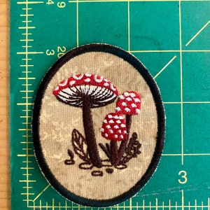 May include: Embroidered patch featuring a cluster of red and white spotted mushrooms with brown stems and a black border. The patch is oval-shaped and set against a beige background with a floral pattern.