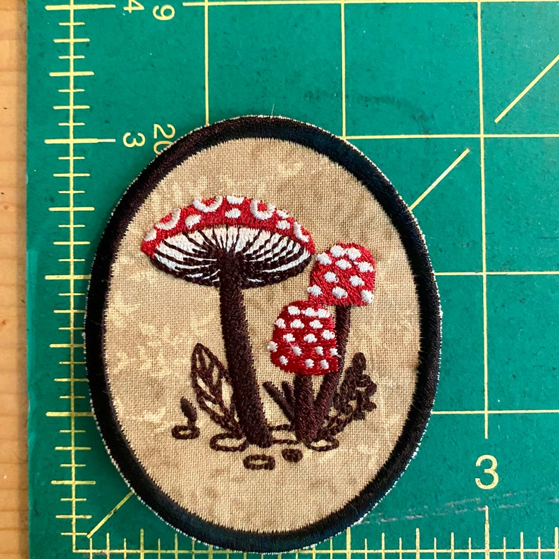 May include: Embroidered patch featuring a cluster of red and white spotted mushrooms with brown stems and a black border. The patch is oval-shaped and set against a beige background with a floral pattern.
