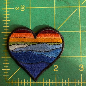 May include: Heart-shaped embroidered patch with a landscape design. The patch features a gradient of colours, including orange, yellow, green, blue, and navy, depicting a sunset over mountains. The patch is outlined in black.