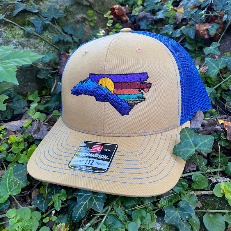 Pode incluir: Bon&eacute; de caminhoneiro bege e azul com um design bordado do contorno do estado da Carolina do Norte, retratando um p&ocirc;r do sol e montanhas. O bon&eacute; tem uma parte traseira de malha azul e uma aba curva com costuras azuis.