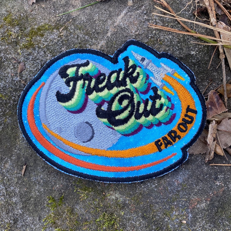 May include: Embroidered patch with the words "Freak Out" in a colorful, stylized font, with a rocket ship blasting off. The patch features a blue border, a gray moon, and the words "Far Out".