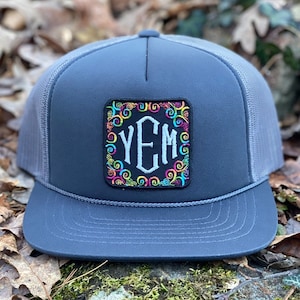 May include: A charcoal gray trucker hat with a mesh back and a flat brim. The front features a square patch with the letters "YEM" in white, surrounded by colorful swirls. A dark gray rope detail accents the brim.