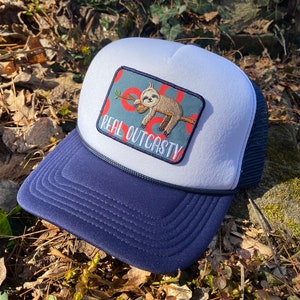 May include: A navy and white trucker hat with a patch showing a sloth clutching a branch. The patch reads "REAL OUTCASTY". The hat has a navy blue peak and a white foam front, with a dark blue mesh back.