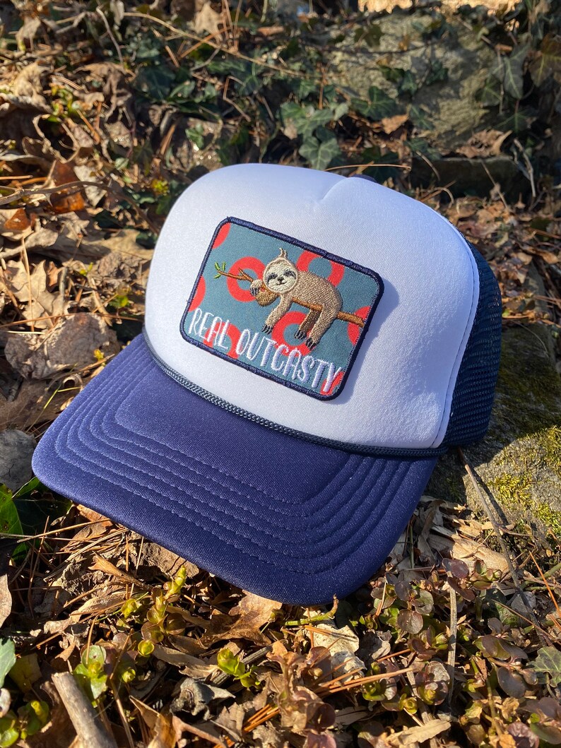 May include: A navy and white trucker hat with a patch showing a sloth clutching a branch. The patch reads "REAL OUTCASTY". The hat has a navy blue peak and a white foam front, with a dark blue mesh back.