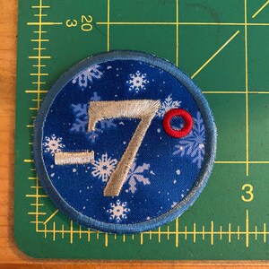 Op de afbeelding: Ronde geborduurde patch met een blauwe achtergrond met witte sneeuwvlokken en het nummer "-7&deg;" in zilveren draad. De patch heeft een blauwe rand en een kleine rode cirkel.