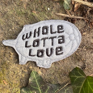 May include: A gray felt patch in the shape of a blimp with the words "WHOLE LOTTA LOVE" embroidered in black. The patch is lying on a bed of dirt and leaves.