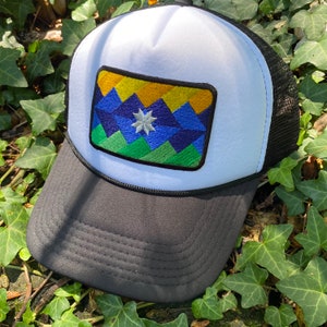 May include: A black and white trucker hat with a mesh back and a curved brim. The front panel is white with a rectangular patch featuring a geometric mountain design in blue, green, yellow, and a white star.
