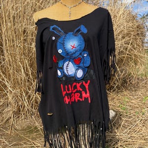 Pode incluir: Top preto com ombros descobertos com bainha desgastada e franjas. A frente apresenta um gráfico de coelho azul com um coração vermelho e as palavras "LUCKY CHARM" em vermelho. Um colar de prata com um pingente também é visível.