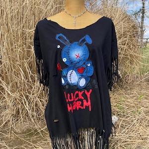 May include: Black distressed t-shirt with a blue bunny graphic and the words "LUCKY HARM" in red. The shirt has fringe detailing along the bottom and sleeves. A silver necklace is visible.