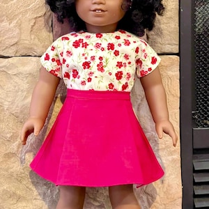 May include: A doll wearing a white top with red floral print and a bright pink skirt. The doll has dark curly hair and a pink bow. The doll is standing in front of a stone wall.