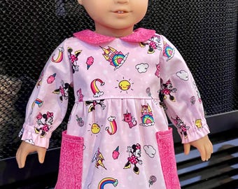 Puede incluir: Una muñeca con un vestido rosa con estampado de Minnie Mouse. El vestido tiene mangas largas, cuello rosa y dos bolsillos rosas. El estampado incluye arcoíris, castillos y soles.