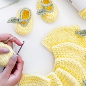 Pode incluir: Conjunto de botinhas e chapéu de bebê de crochê, em fio amarelo macio. As botinhas apresentam um design Mary Jane com um detalhe de folha verde. Uma mão está fazendo crochê em uma parte do conjunto com uma agulha de crochê.