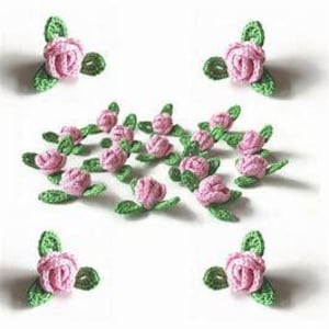 Puede incluir: Rosas rosas de ganchillo con hojas verdes. Los adornos florales hechos a mano están dispuestos sobre una superficie blanca. Las rosas son de color rosa suave y las hojas de un verde vibrante.