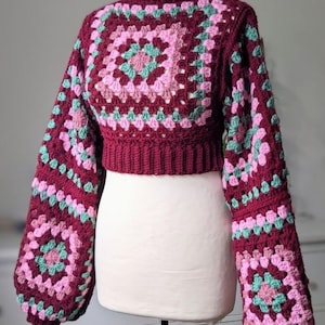 Puede incluir: Un suéter de ganchillo burdeos con cuello alto y mangas largas y abullonadas. El suéter presenta un patrón de punto de abuela cuadrado en tonos burdeos, rosa y verde azulado. El diseño recortado se muestra en un maniquí blanco.
