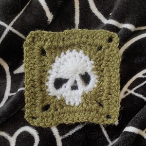 May include: A square, crocheted patch featuring a white skull design centered on an olive green background. The skull's eye sockets and nose are black. The patch is made with a textured stitch.
