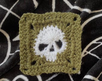 May include: A square, crocheted patch featuring a white skull design centered on an olive green background. The skull's eye sockets and nose are black. The patch is made with a textured stitch.