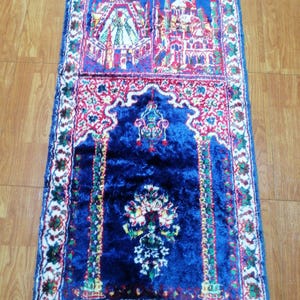 Könnte beinhalten: Ein rechteckiger Gebetsteppich mit einem tiefblauen Mittelfeld und einem detaillierten Rand. Der obere Bereich zeigt Illustrationen von Gebäuden in Rot, Weiß und Blau. Der Teppich hat einen Fransenrand und komplizierte Blumenmuster.