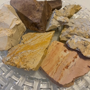 Könnte beinhalten: Eine Sammlung roher, unraffinierter Tonstücke in verschiedenen Erdtönen wie Creme, Goldgelb, Hellbraun und Altrosa. Die Stücke weisen unregelmäßige, gezackte Kanten und strukturierte Oberflächen auf und sind auf einem klaren Glasteller angerichtet.
