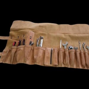 Puede incluir: Un rollo de herramientas de cuero marrón con múltiples bolsillos y correas. Varias herramientas, incluyendo llaves, destornilladores y alicates, son visibles en los bolsillos. El rollo está diseñado para mantener las herramientas organizadas y accesibles.