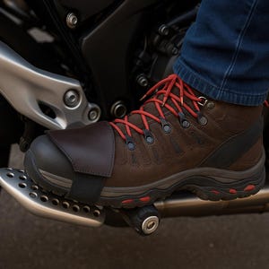 Może przedstawiać: Zbliżenie brązowego skórzanego buta motocyklowego z czerwonymi sznurowadłami i czarną podeszwą. But jest na podnóżku motocykla. But ma ciemnobrązową skórzaną osłonę. Motocykl jest czarny i srebrny.