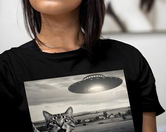 May include: Black t-shirt featuring a grayscale image of a cat taking a selfie with a UFO in the background. The cat has a surprised expression. The UFO is a classic flying saucer shape.