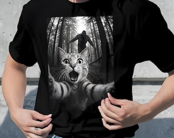 May include: Black t-shirt featuring a black and white graphic of a surprised cat taking a selfie in a forest, with a Bigfoot figure in the background. The cat has wide eyes and an open mouth.