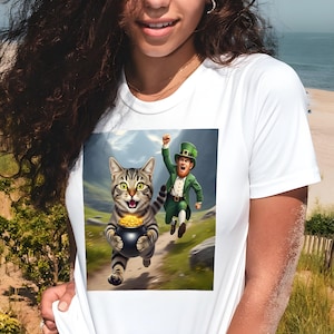Pode incluir: Camiseta branca com um desenho de um gato malhado correndo com um pote de ouro, e um duende correndo atrás. O gato tem olhos verdes e está correndo em um caminho. O duende está usando um terno e chapéu verdes.