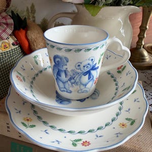 May include: A white porcelain teacup and saucer set with a blue floral and teddy bear design. The cup has a heart-shaped handle. The set includes a teacup, a small plate, and a larger plate, all with scalloped edges.