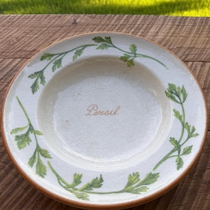 May include: A round ceramic plate with a cream-colored base featuring a fine crackle glaze finish. The rim is decorated with green painted parsley sprigs, and the word Persil is written in the center in a brown cursive script font.