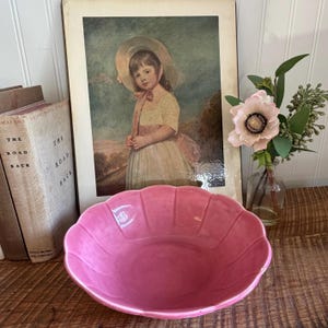 May include: A glossy, scalloped-edge pink ceramic bowl with a rounded basin. The bowl features a smooth, reflective surface and a deep, vibrant pink color, positioned in front of a framed portrait of a child and a glass vase with a pink flower.