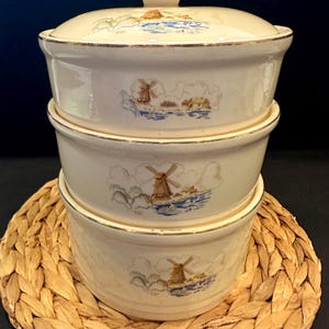 Puede incluir: Juego de fuentes de cerámica apiladas, de color crema, con tapas. Cada fuente presenta una escena de molino de viento en azul, marrón y beige. Los bordes están adornados con dorado. Las fuentes están sobre un salvamanteles tejido.