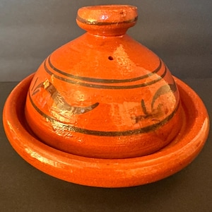 May include: A vibrant orange ceramic tagine pot with a conical lid and a circular base. The lid features black decorative lines and a small knob. The tagine is designed for cooking and serving Moroccan dishes.