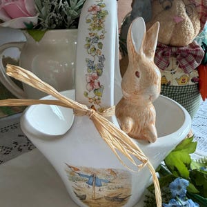 Puede incluir: Cesta de cerámica blanca con asa decorada con motivos florales y un lazo de paja. La cesta presenta una escena pintada con un espantapájaros y pájaros. Una pequeña figura de conejito de cerámica está dentro.