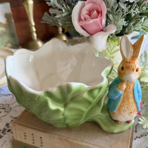 Può includere: Un vaso di ceramica a forma di foglia di cavolo, con una statuina di Peter Coniglio. Il vaso è verde chiaro con l'interno bianco. Peter Coniglio indossa una giacca blu. Fiori sullo sfondo.