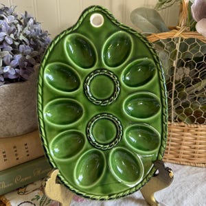 Puede incluir: Una bandeja ovalada de cerámica verde brillante para servir huevos, con doce hendiduras en forma de huevo dispuestas en un patrón simétrico. La bandeja tiene un borde decorativo estilo cuerda y un orificio en la parte superior para colgar.